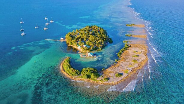 Vue aérienne d’une île tropicale aux eaux turquoise et limpides, avec un petit village composé de quelques bâtiments et plusieurs voiliers ancrés à proximité.
