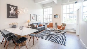 Een moderne woonkamer met een bank, salontafel en stoelen. Er is een groot raam, kunst aan de muur en een Louis Vuitton bord.