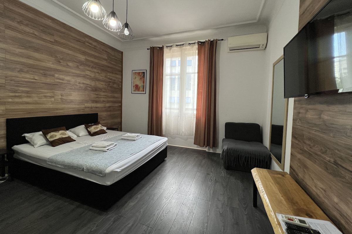 A modern hotel room with a wooden headboard, a large bed with a neatly made bed, a desk with a chair, a flat-screen TV, and a window with curtains.