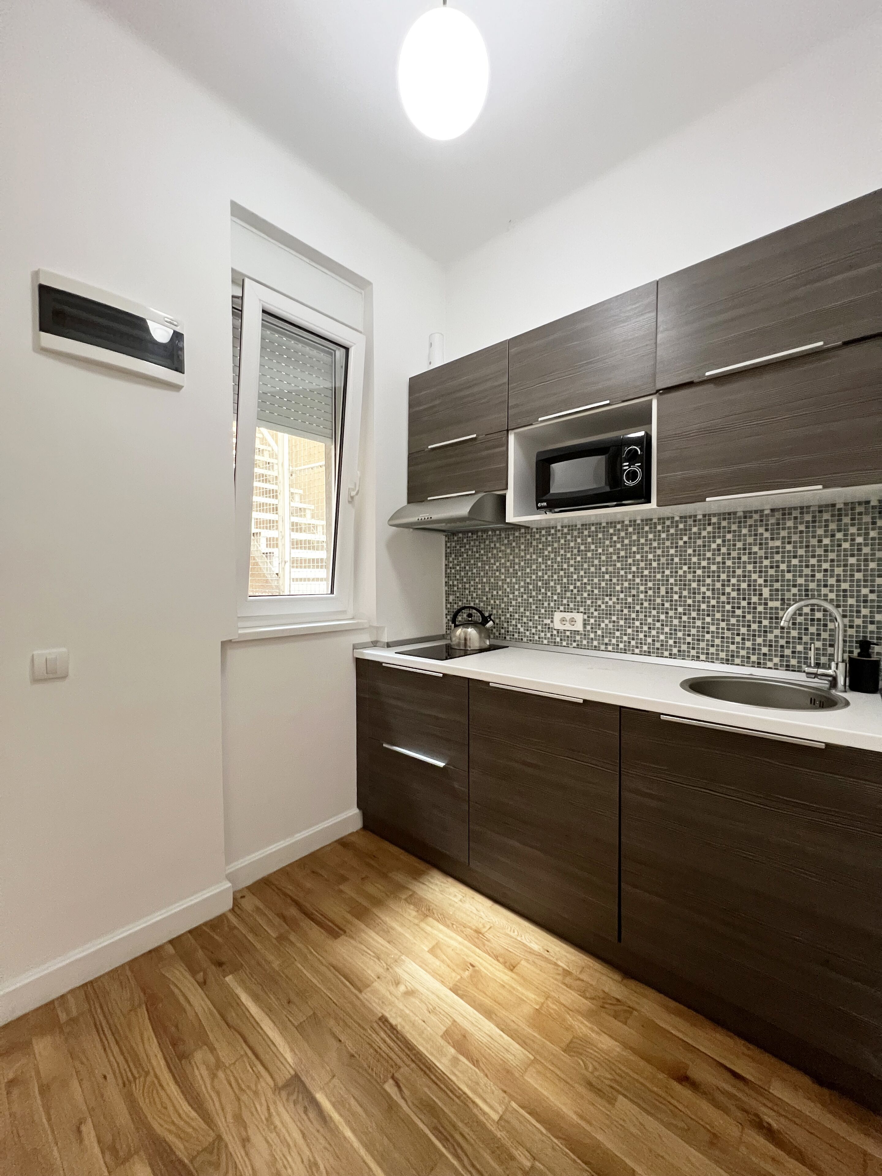 Una cocina compacta con armarios de madera oscura, una encimera blanca, un microondas y un salpicadero de azulejos.