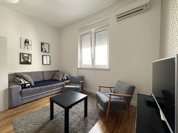 Een woonkamer met een grijze bank, twee fauteuils, een zwarte salontafel en een tv-meubel. Er hangen foto's in lijsten aan de muur en er is een raam met jaloezieën.