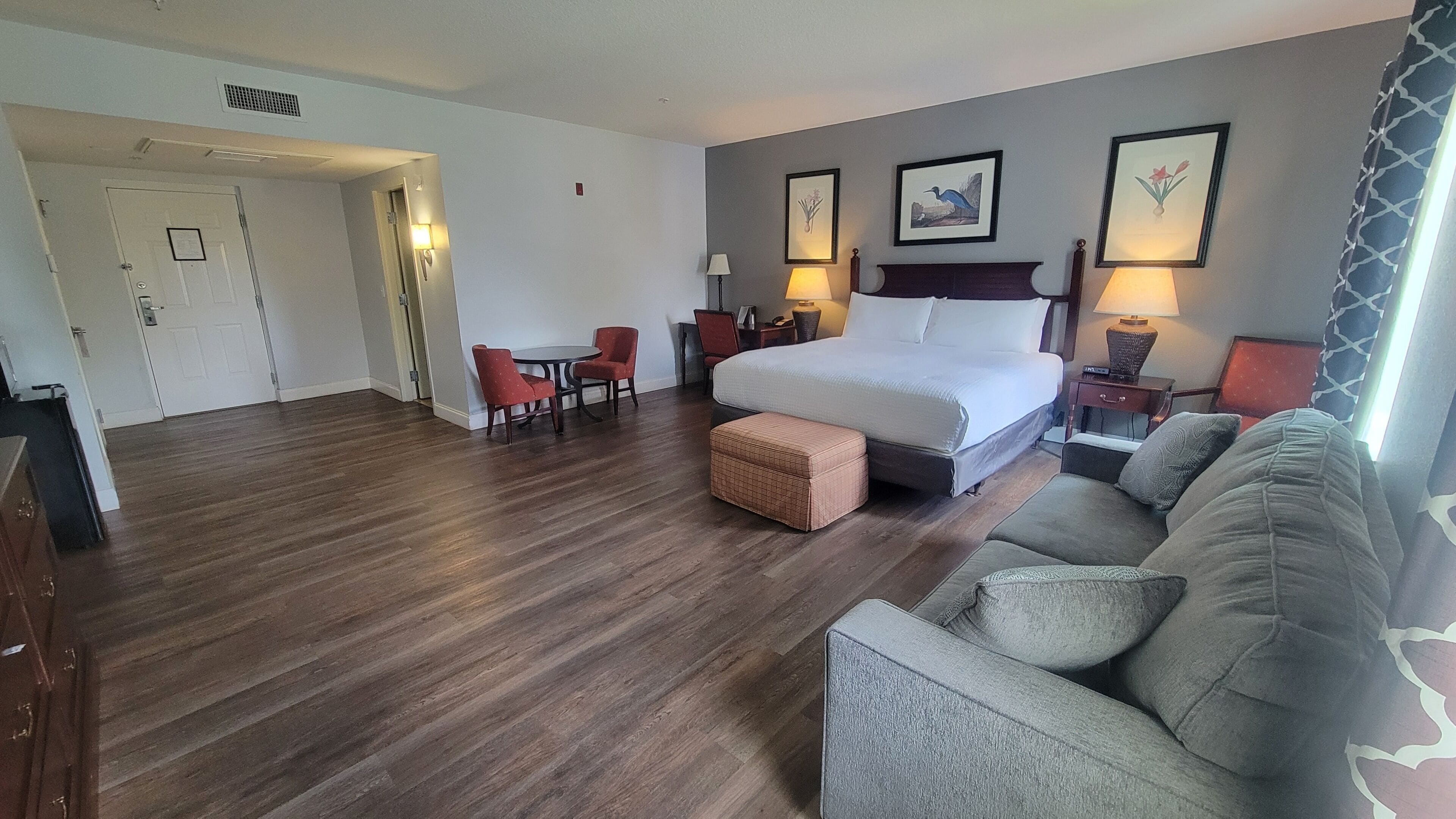 Quarto de hotel com cama, sofá, poltrona, mesa pequena e luminária.