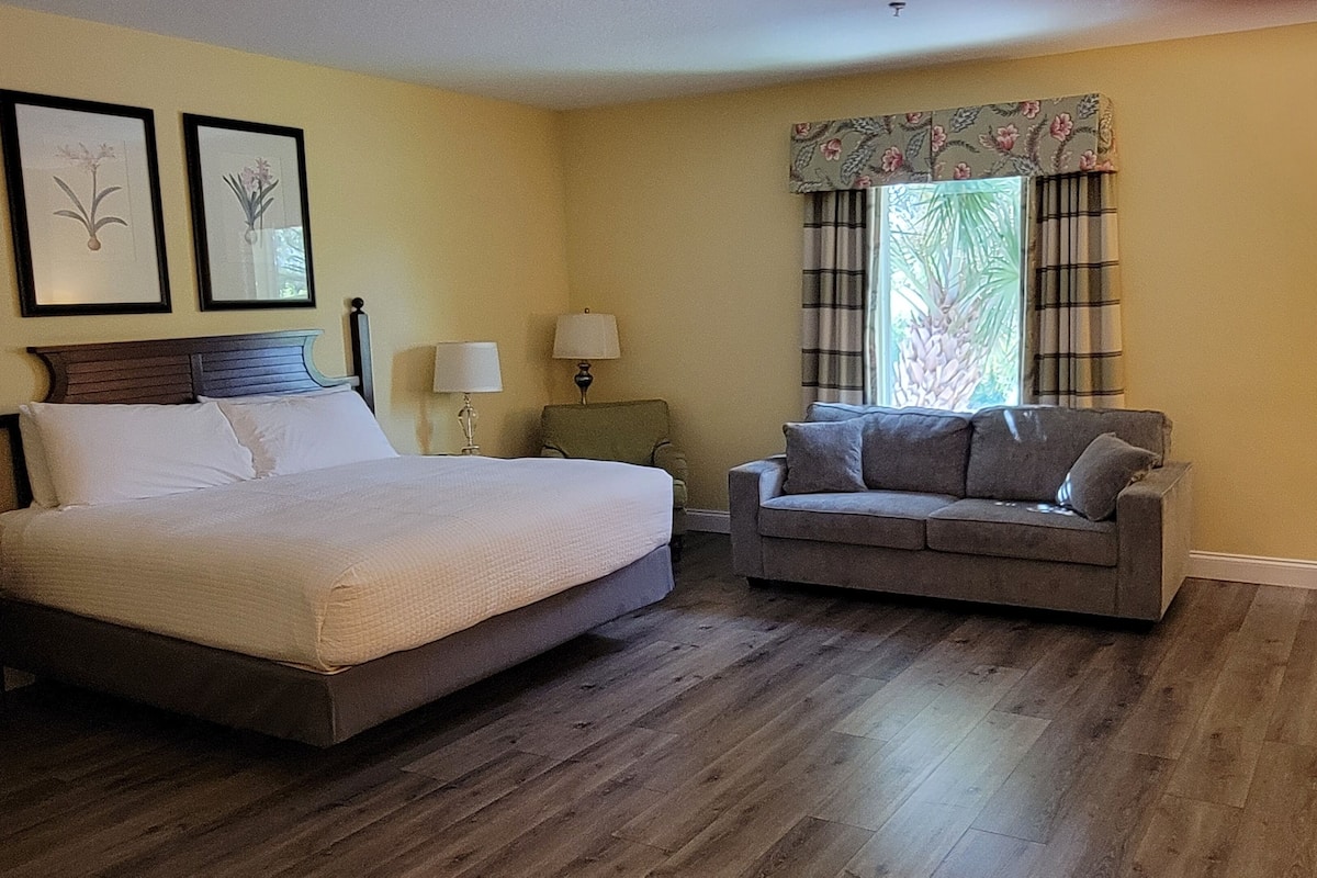 Habitación de hotel con cama, sofá, sillón, mesa, lámpara, ventana con cortinas y dos cuadros en la pared.