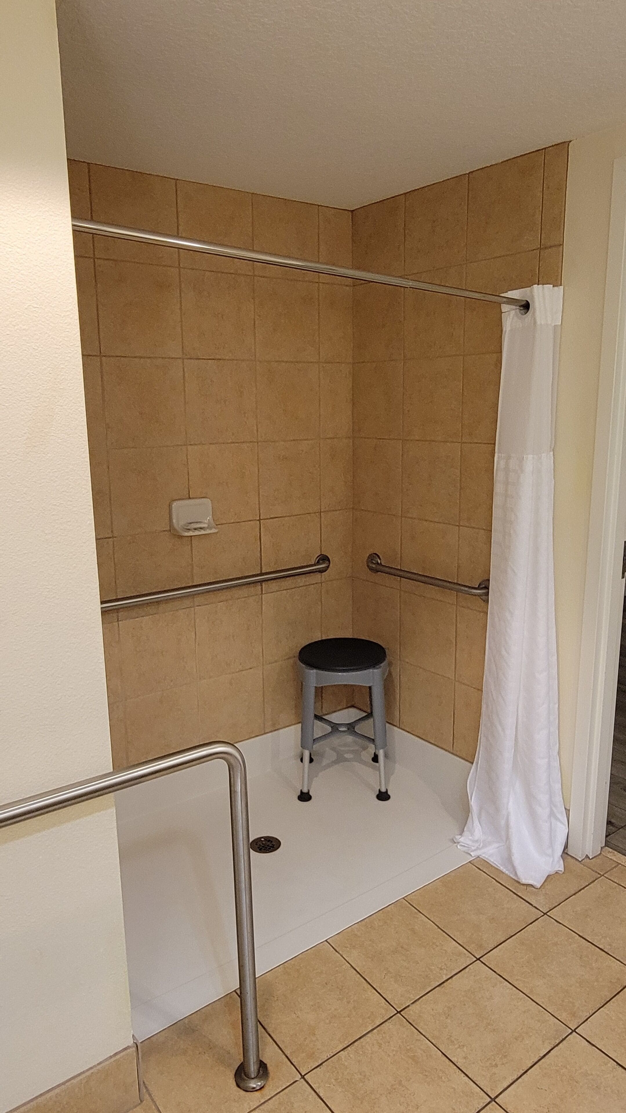 A small, enclosed shower area with a white shower curtain, a grab bar, and a step stool.