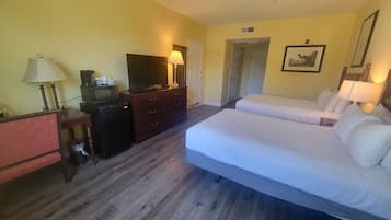 A hotel room with a bed, a desk with a lamp, a TV, a dresser, and a door leading to another room.