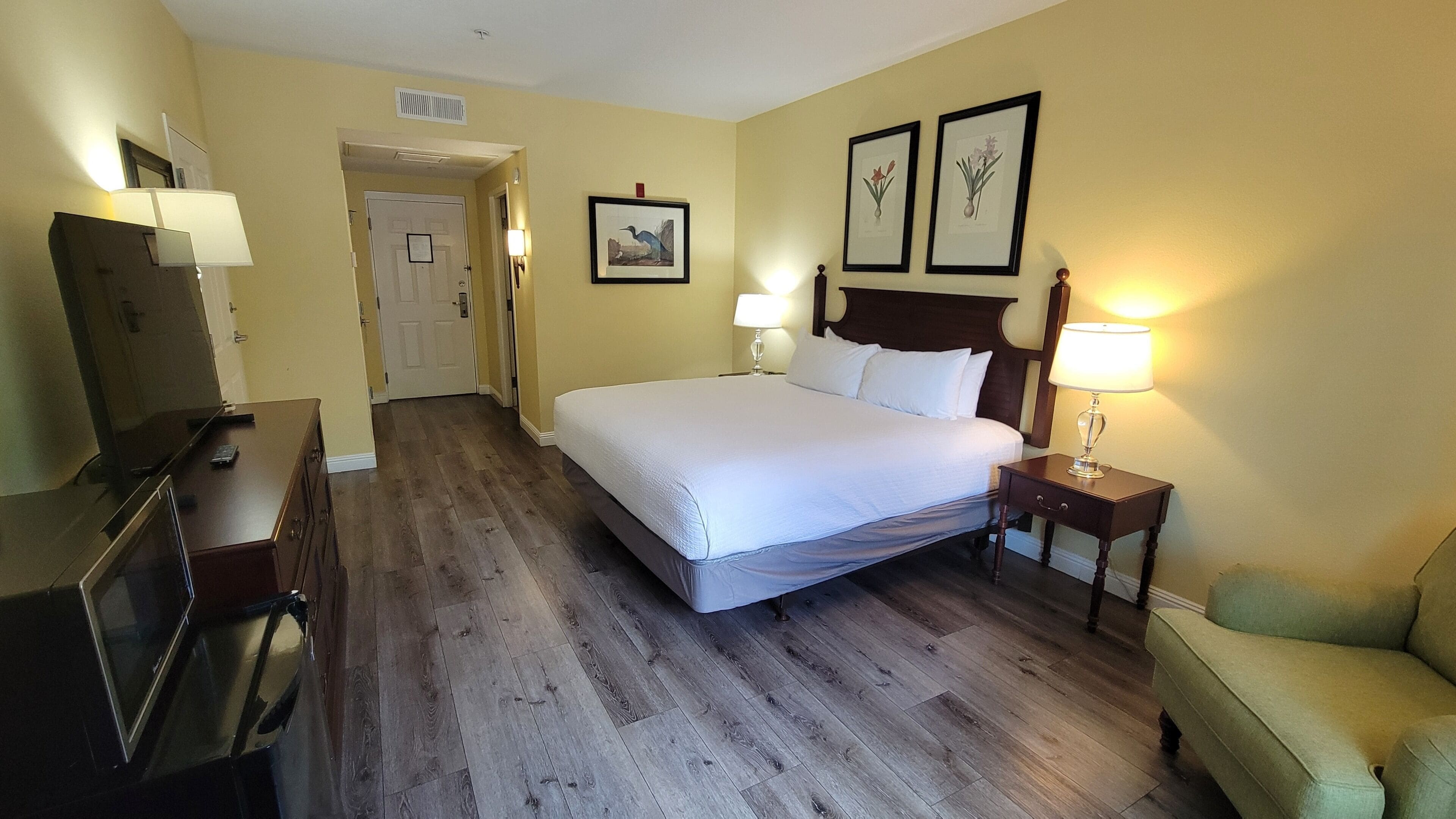 Ein Hotelzimmer mit Bett, Nachttischlampen, Fernseher, Kommode und gerahmten Bildern an der Wand.