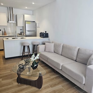 Una sala de estar moderna con un sofá, una mesa de centro de vidrio y una zona de cocina con refrigerador y microondas.
