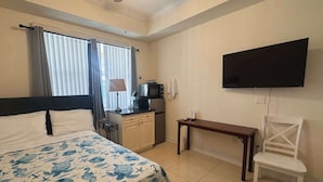 A hotel room with a bed, a desk, a chair, a TV, a kitchenette, and a window with curtains.