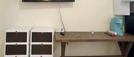 Ein Zimmer mit einem an der Wand befestigten Fernseher, zwei weißen Stauraumeinheiten mit braunen Griffen, einem Holztisch und einer Kaffeemaschine.