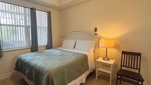 A bedroom with a bed, a chair, a nightstand with a lamp, a window with blinds, and a wall-mounted phone. - Bright Studio – Free WiFi – Bonita Springs (Bonita Springs)