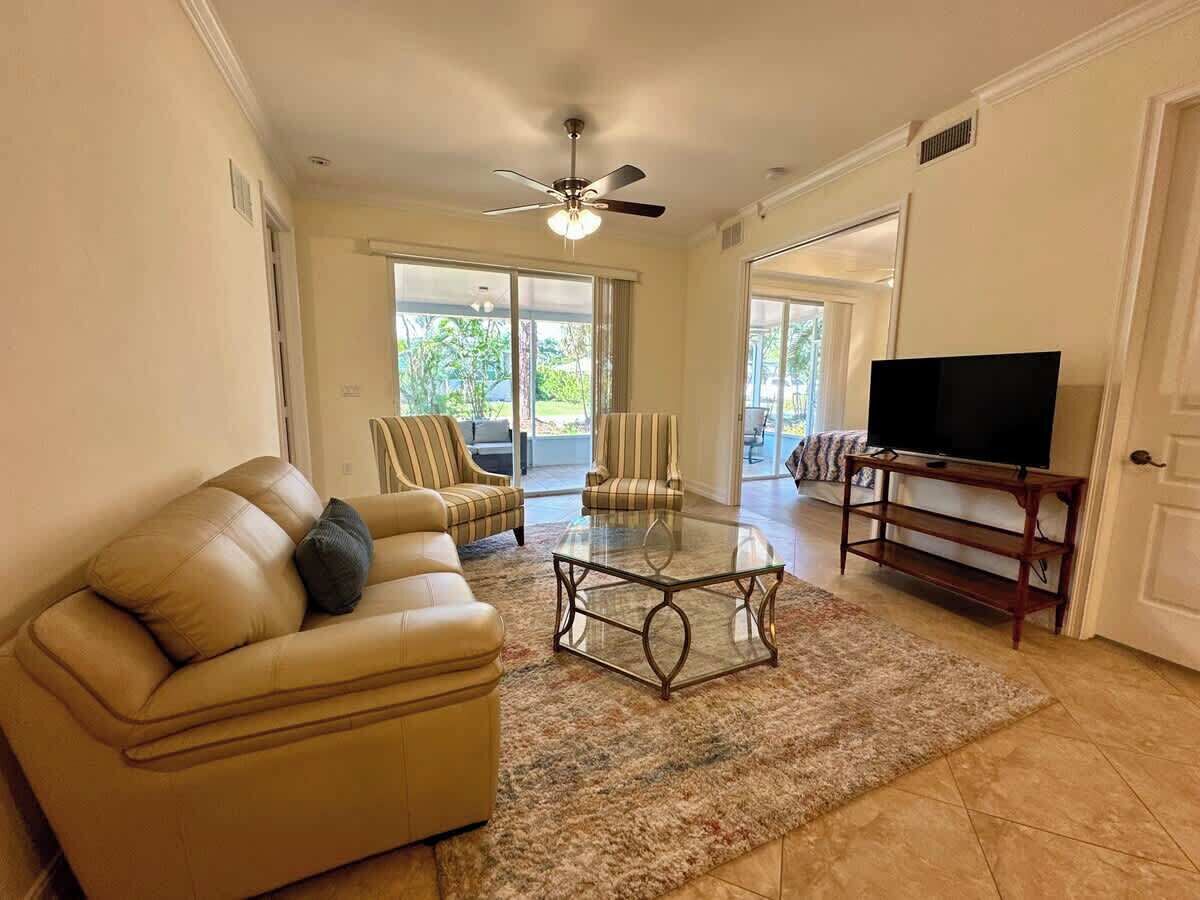 A living room with a beige sofa, a glass coffee table, striped armchairs, a TV on a wooden stand, and a ceiling fan.