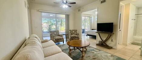 A living room with a beige sofa, a wooden coffee table, and a television on a stand. There are two armchairs and a patterned rug.