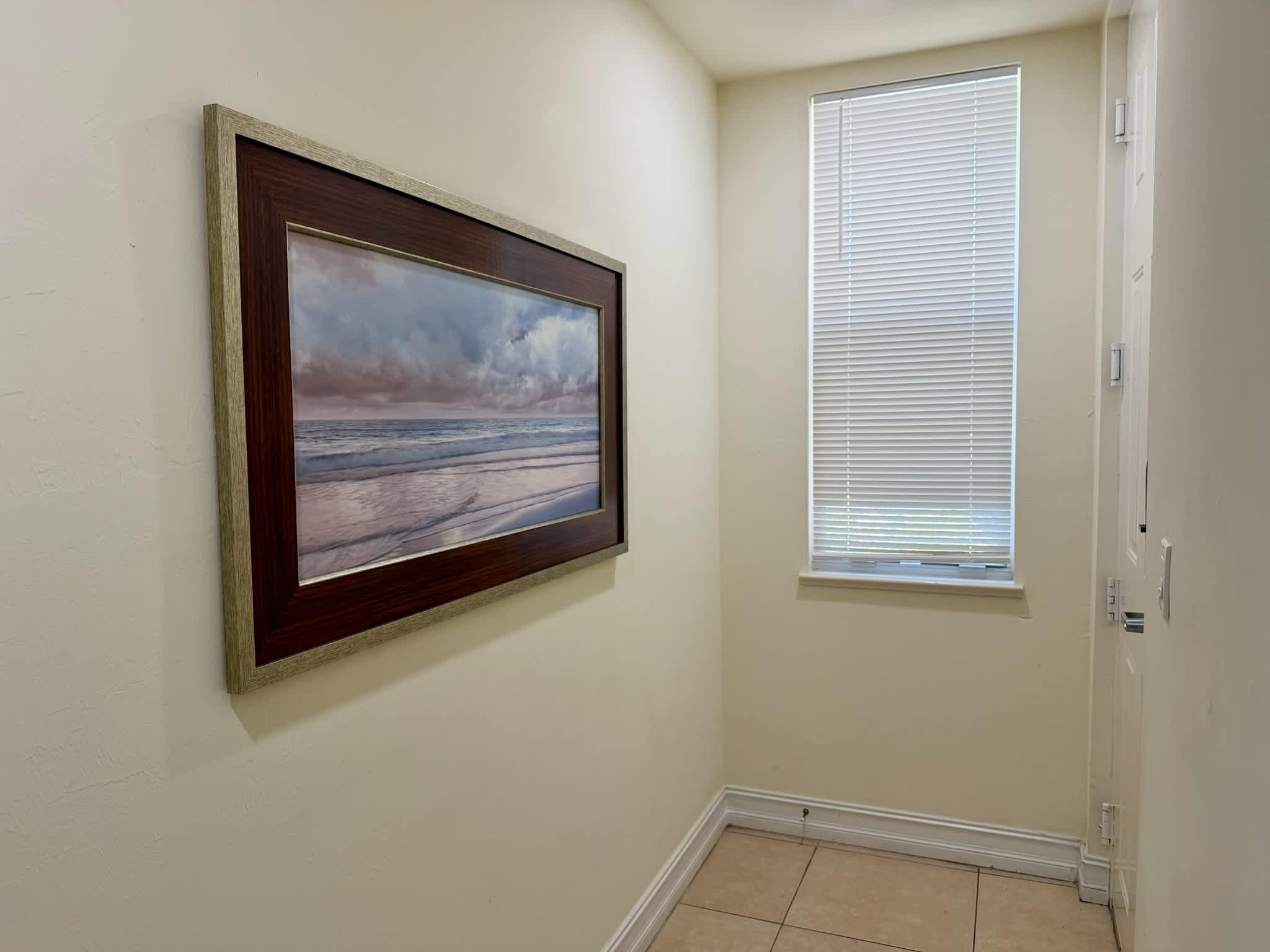 A room with a framed painting of a beach scene on the wall, a window with blinds, and a tiled floor.