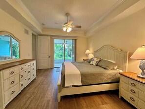 Ein Schlafzimmer mit Bett, Nachttischen, Kommode, Deckenventilator und Blick durch eine Glastür.