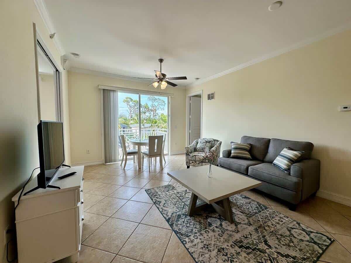 A living room with a grey sofa, a coffee table, a TV on a cabinet, and a balcony with a view of trees.