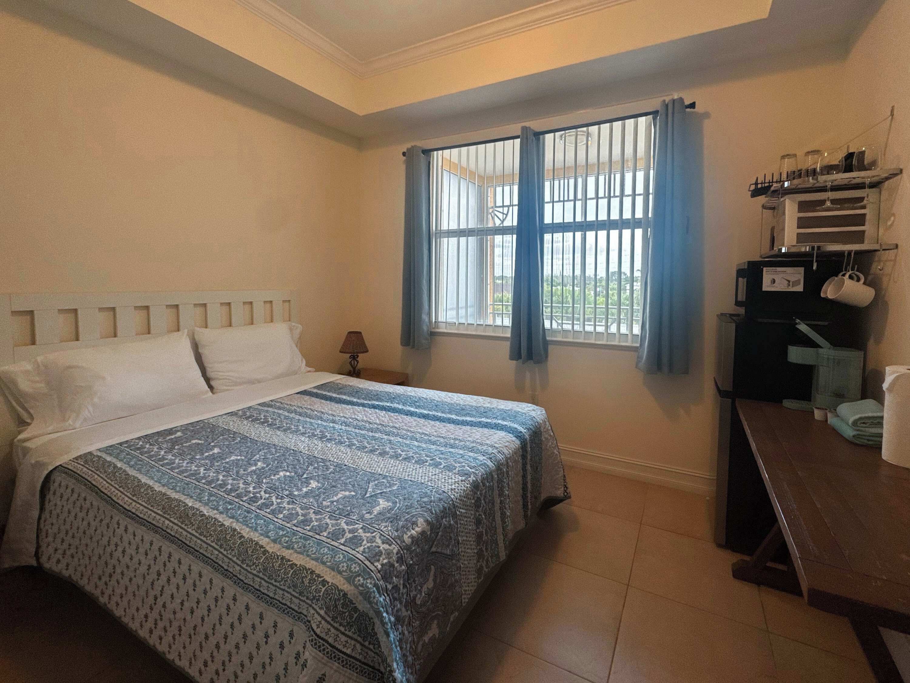 Une chambre d’hôtel comprenant un lit, une table de chevet, une fenêtre avec des rideaux et un coin cuisine équipé d’un four à micro-ondes et d’une machine à café.