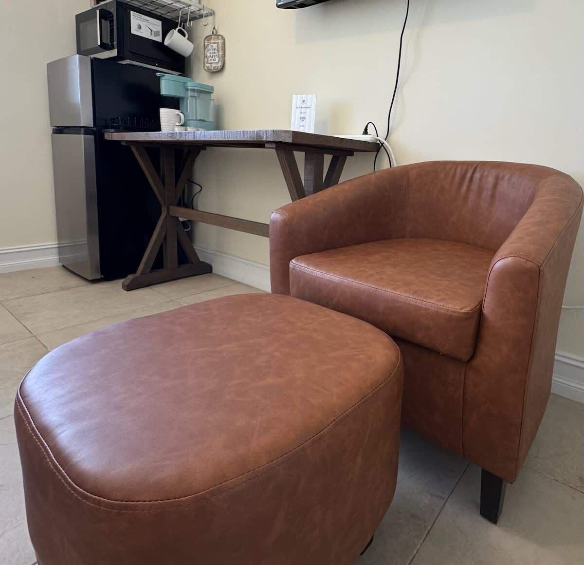 A brown leather armchair and ottoman in a room with a kitchenette area.