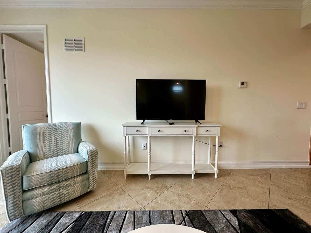 Una sala de estar con un sofá, un televisor sobre un mueble blanco y piso de baldosas.