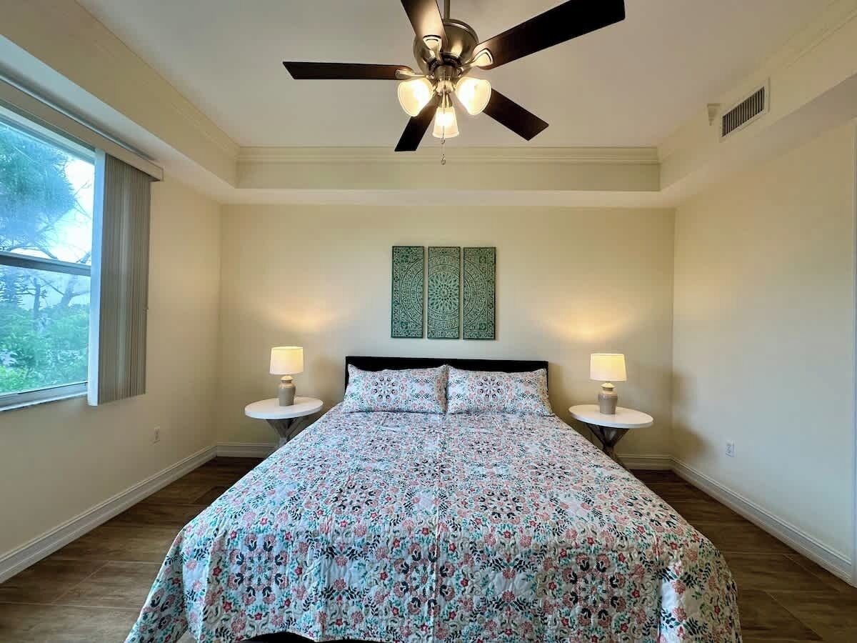 A bedroom with a bed, two bedside tables with lamps, a ceiling fan, and a window with curtains.