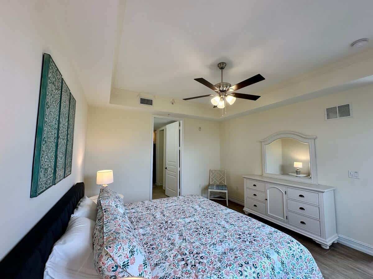 A bedroom with a bed, a dresser, a ceiling fan, and a door leading to another room.