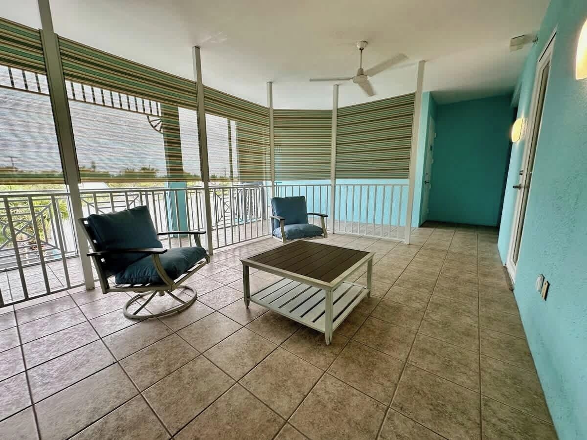 A covered balcony with tiled flooring, two chairs, a table, and a ceiling fan.