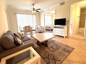 Ein Wohnzimmer mit Sofa, Couchtisch, Fernseher, Deckenventilator und Blick ins Badezimmer.