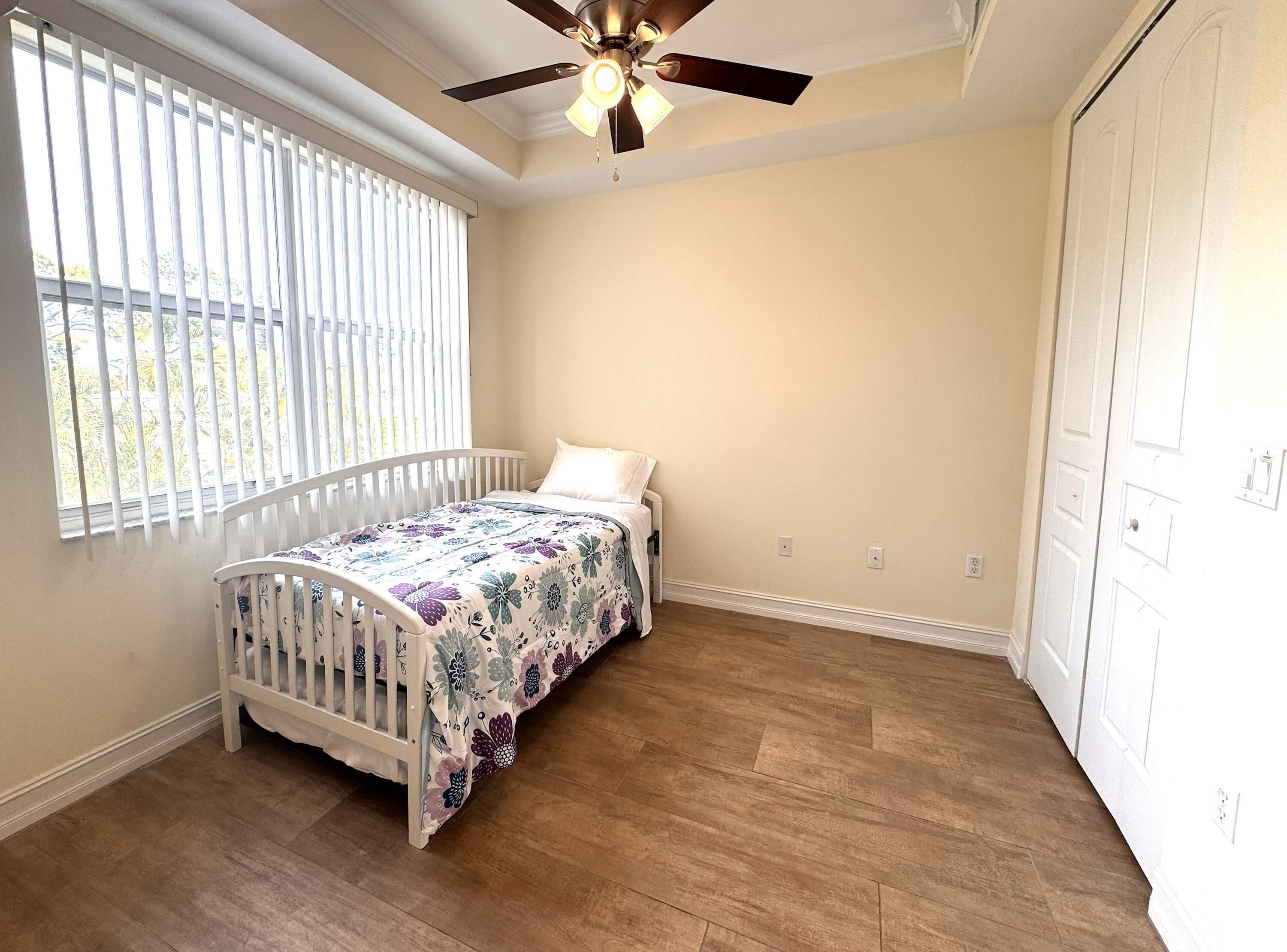 A bedroom with a single bed, a ceiling fan, a window with blinds, and a wooden floor.