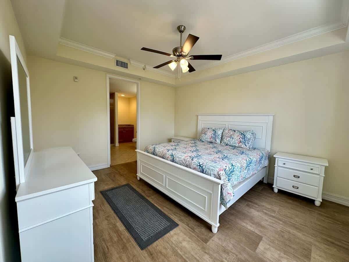Une chambre à coucher comprenant un lit, des tables de chevet, une commode, un ventilateur de plafond et une porte donnant sur une autre pièce.