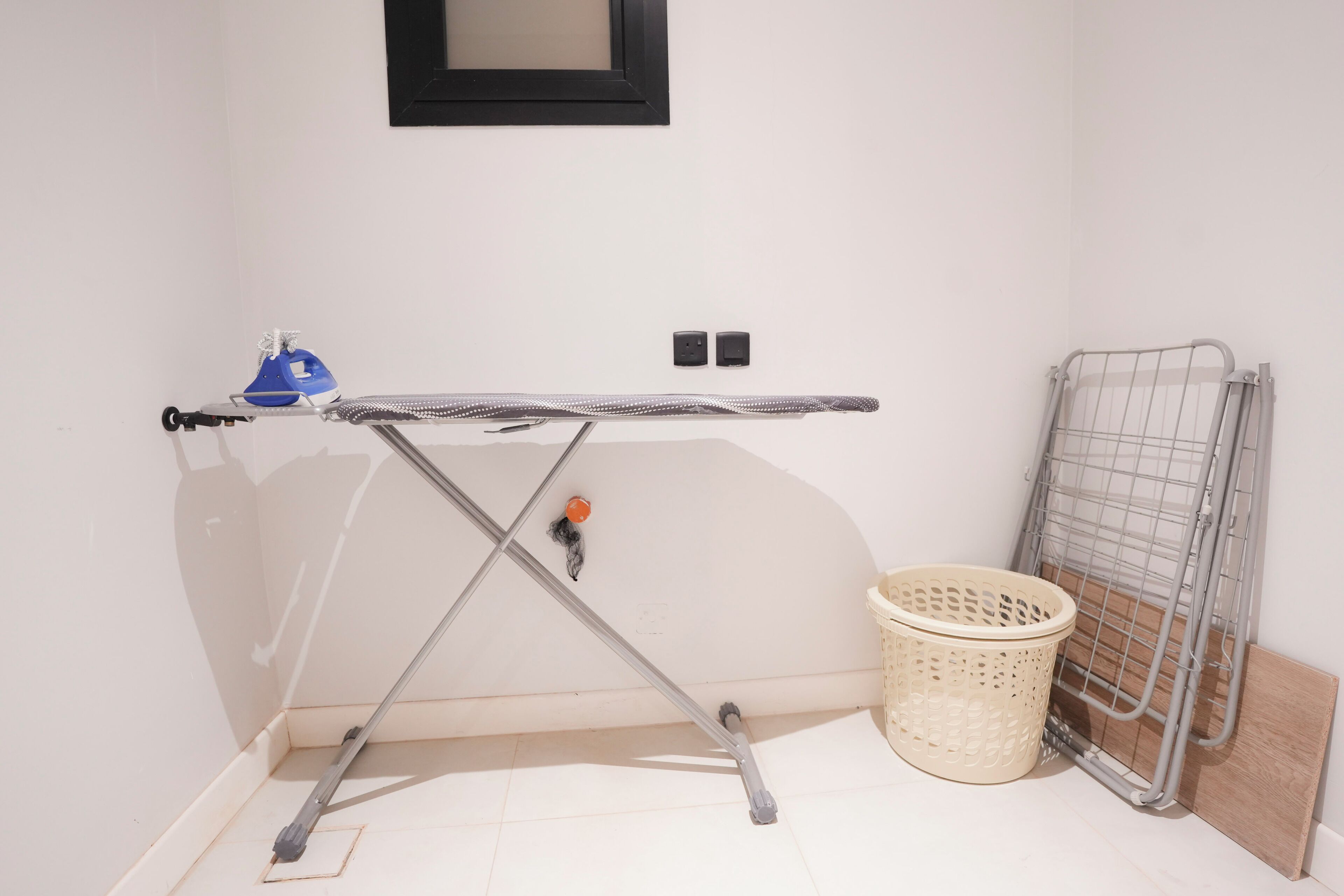 Un coin avec une planche à repasser en métal, un panier à linge et un étendoir en métal.