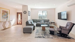Un salon moderne avec un canapé d’angle, un pouf et un fauteuil inclinable. Un téléviseur à écran plat est fixé au mur, et une petite table d’appoint est placée à proximité.