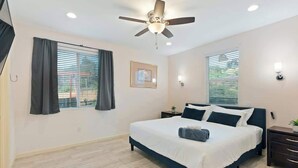 Une chambre à coucher comprenant un lit, un ventilateur de plafond, une fenêtre avec des stores, une table de chevet et une lampe fixée au mur.
