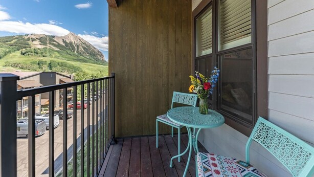 Un balcon doté d’un plancher en bois, deux chaises turquoise, une petite table avec un vase de fleurs et une vue sur une montagne et un parking.