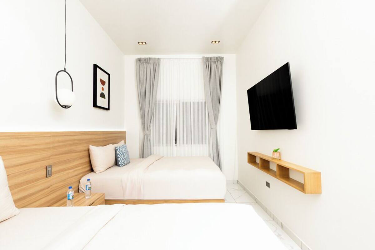 Un dormitorio moderno con cabecera de madera, cama con ropa blanca, televisor montado y un pequeño estante de madera con una planta.