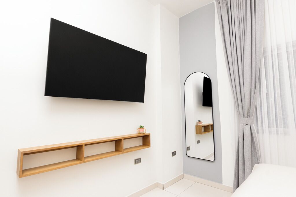 A modern bedroom with a mounted TV, a mirror, and a wooden shelf.
