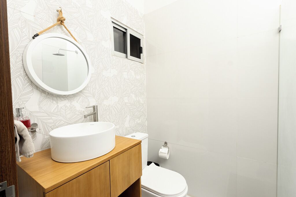 A bathroom with a round mirror, a white sink, wooden cabinets, a toilet, and a shower area.