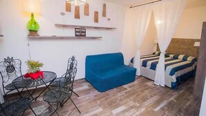 Une petite pièce agréable, avec un canapé bleu, un lit à baldaquin, une table entourée de chaises et une étagère fixée au mur, ornée d’objets décoratifs.