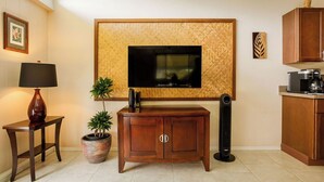 A living room with a flat-screen TV mounted on a textured wall, a wooden cabinet, a potted plant, a standing fan, and a coffee machine.