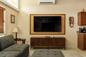 A living room with a flat-screen TV mounted on the wall, a wooden cabinet below, a sofa, a lamp, and a window with blinds. - Now & Zen Retreat (Hilo)
