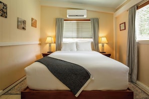 A hotel room with a large bed, two bedside tables with lamps, a window with blinds, and an air conditioning unit.