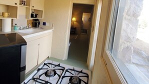 Une cuisine de taille réduite, équipée d’une cuisinière à gaz, d’un évier et de placards. Une fenêtre se trouve sur le côté droit.