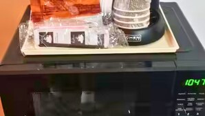 A coffee maker on a microwave with a timer set to 1047, a plastic container, and a wrapped item. - OSU 223 Queen Bed Hotel Room (Stillwater)