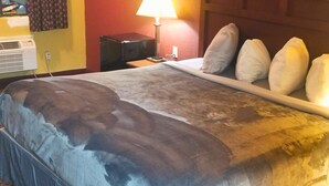A hotel room with a bed, a nightstand, a lamp, and a wall-mounted air conditioner. - OSU 223 Queen Bed Hotel Room (Stillwater)