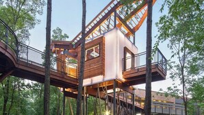 Una casa en un árbol con fachada de madera, barandales metálicos y una escalera que lleva hasta ella.