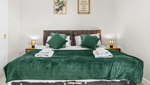 A neatly made bed with a green blanket, two pillows, and two folded towels. There are two wall-mounted picture frames, one with a plant and the other with text. - Modern | Chester Retreat | Free Parking | Sleeps 6 | (Chester)