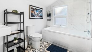 A modern bathroom with a white toilet, a white bathtub, a black shelving unit with plants, a framed abstract painting, and a patterned floor. - Modern | Chester Retreat | Free Parking | Sleeps 6 | (Chester)