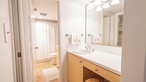 Baño con lavamanos blanco, espejo y toallero. Se observa un inodoro al fondo.