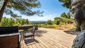 A wooden deck with wicker furniture and a statue, overlooking a scenic landscape with trees and distant hills. - Maison piscine proche Alpilles (Noves)