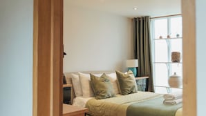 A bedroom with a wooden door frame, a bed with green bedding, a bedside table with a lamp, and a window with curtains. - Bull Pen Cottage | A Luxury Cosy Countryside Retreat (Beaconsfield)