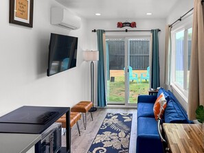 A living room with a sofa, a TV, a dining table, and a sliding glass door leading to an outdoor area. - Hibiscus Hale - Perfect for 2! (Waialua)
