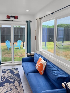 A room with a blue sofa, a patterned rug, a large window, and a sliding door leading to an outdoor area with chairs and a fence. - Hibiscus Hale - Perfect for 2! (Waialua)